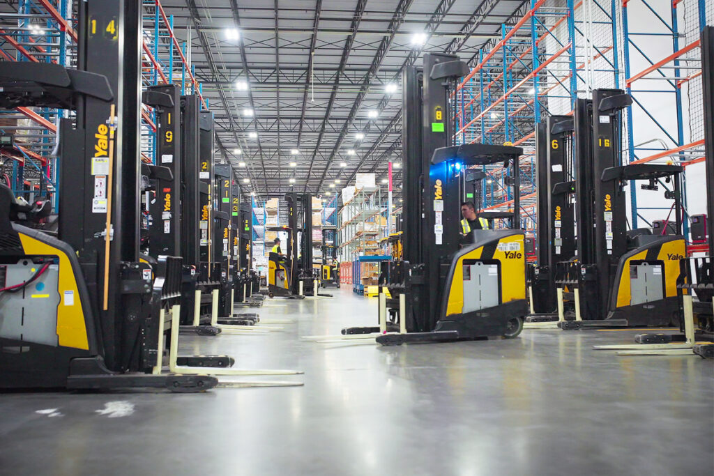Busy warehouse/3PL with lithium ion battery electric standup reach trucks (forklifts) and shelving.